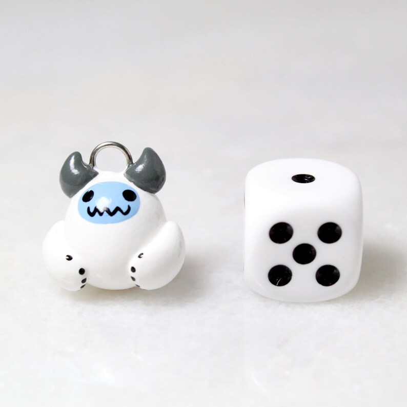 A  yeti charm next to a board game die. The charm is about the same size as the dice.