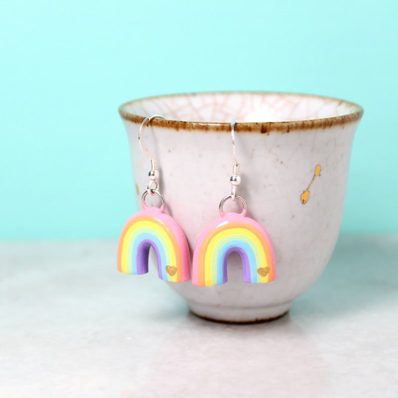 A pair of pastel rainbow arch earrings with tiny golden hearts. The earrings are hanging from a small cup in front of a bright blue background.