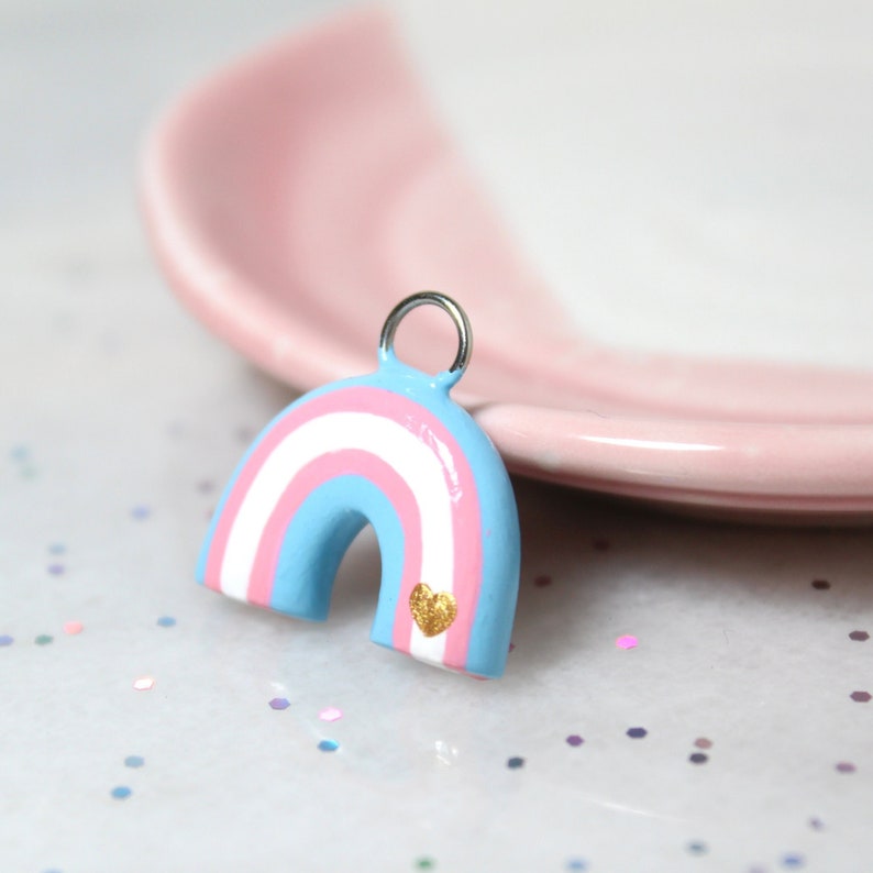 A rainbow arch charm with the colors of the trans pride flag (blue, pink, and white) with a tiny golden heart. The charm is resting against a pink jewelry dish.