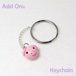 May include: A pink pig-shaped keychain charm with black eyes and a small nose, attached to a silver chain and keyring. The text "Add Ons:" and "Keychain" are visible in the image.