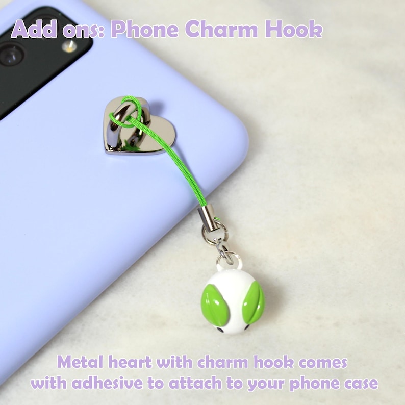 May include: A silver metal heart phone charm hook with a green cord and a white and green charm. The text "Add ons: Phone Charm Hook" and "Metal heart with charm hook comes with adhesive to attach to your phone case" are visible.