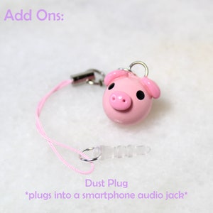 May include: Pink pig charm with black eyes and a pink nose. The charm is attached to a pink string with a clear plastic dust plug. The dust plug is designed to fit into a smartphone audio jack. The text "Add Ons:" and "Dust Plug *plugs into a smartphone audio jack*" is visible in the image.