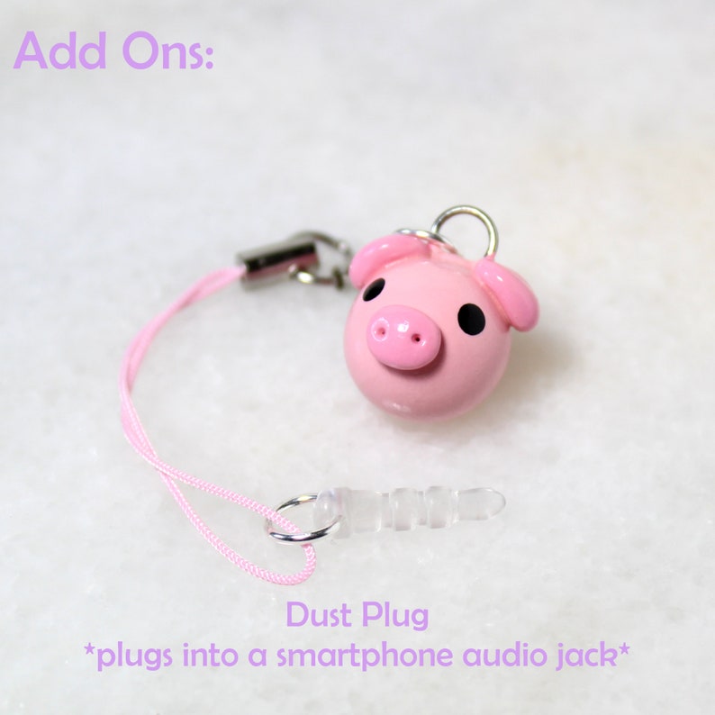 May include: Pink pig charm with black eyes and a pink nose. The charm is attached to a pink string with a clear plastic dust plug. The dust plug is designed to fit into a smartphone audio jack. The text "Add Ons:" and "Dust Plug *plugs into a smartphone audio jack*" is visible in the image.
