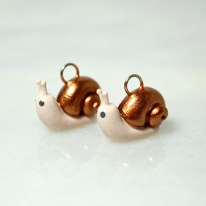May include: Two snail-shaped charms. The snail bodies are a pale pink, with metallic copper-colored shells. Each charm has a small silver loop at the top for hanging. The snails have small black eyes and antennae.