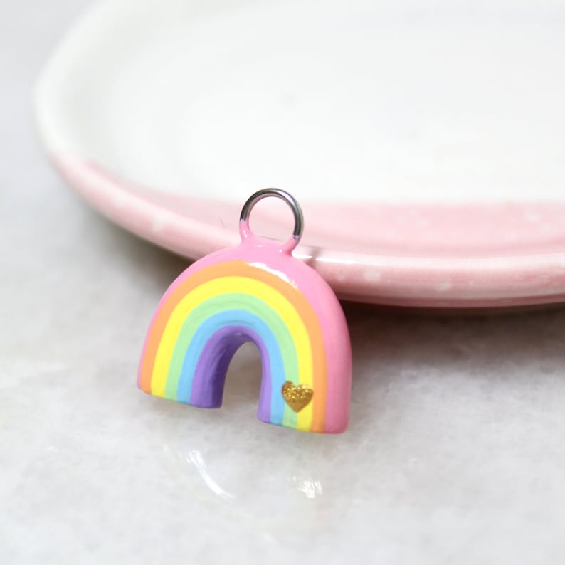 A pastel rainbow arch charm with a tiny gold heart resting against a jewelry dish.