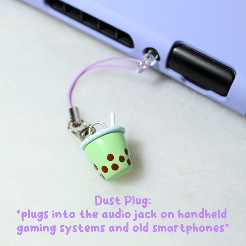 May include: A green and brown boba tea cup dust plug with a light blue rim and a white straw. The dust plug is attached to a purple cord and silver chain. The text "Dust Plug" and a description are below the dust plug.