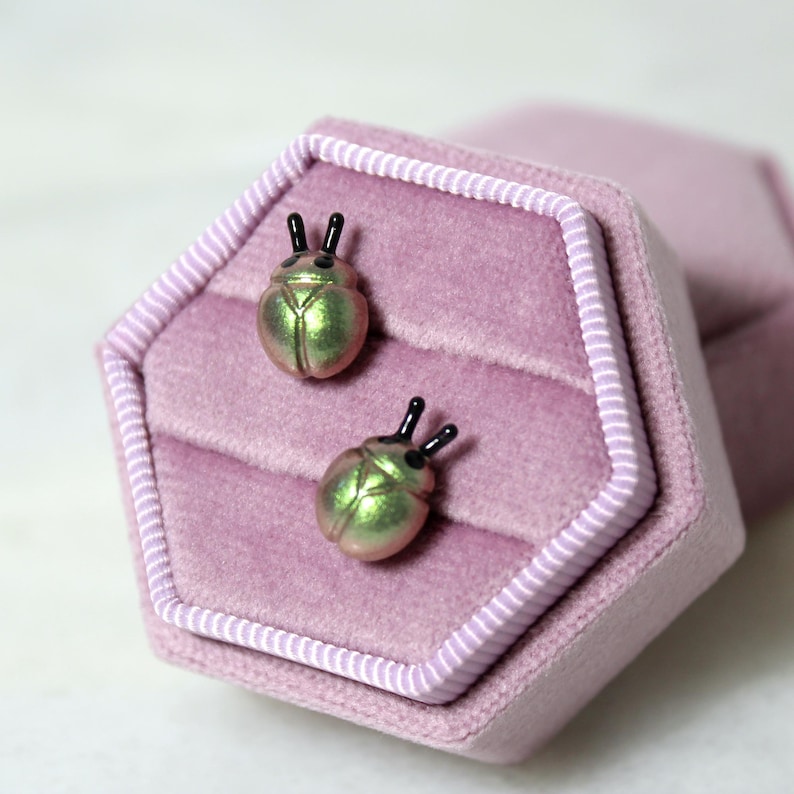 May include: A pair of iridescent green and pink beetle-shaped stud earrings with black antennae, presented in a pink velvet jewelry box. The earrings have a metallic sheen and are set against a soft, textured background.