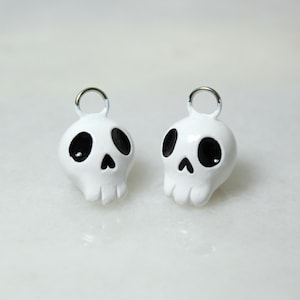 May include: Two white skull-shaped charms with black eye sockets and a nose. Each charm has a silver metal loop at the top. The skulls have a glossy finish and are set against a light background.
