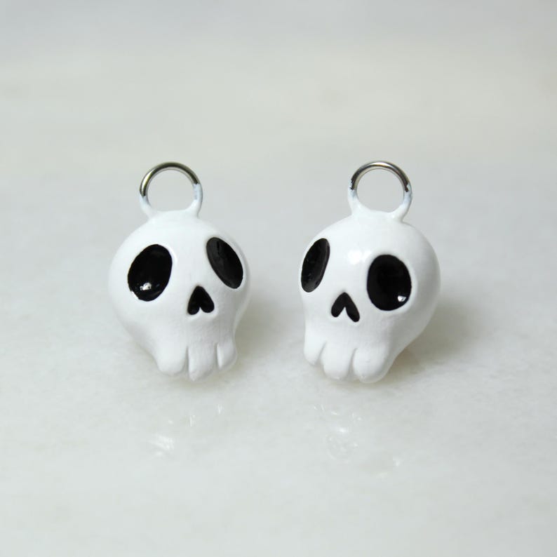 May include: Two white skull-shaped charms with black eye sockets and a nose. Each charm has a silver metal loop at the top. The skulls have a glossy finish and are set against a light background.