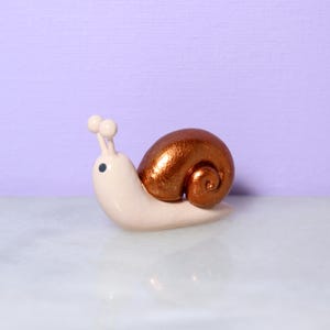 A miniature snail figurine with a sparkly bronze shell sits against a light purple background