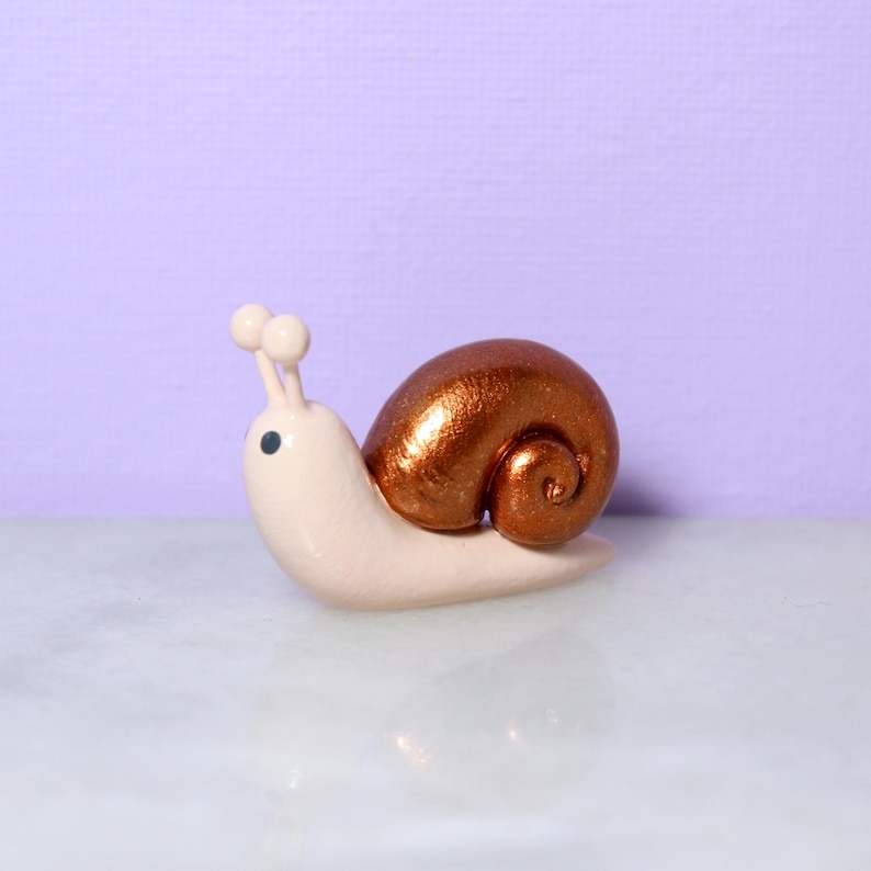 A miniature snail figurine with a sparkly bronze shell sits against a light purple background