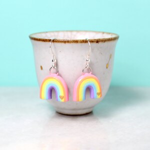 A pair of pastel rainbow arch earrings with tiny golden hearts. The earrings are hanging from a small cup in front of a bright blue background.