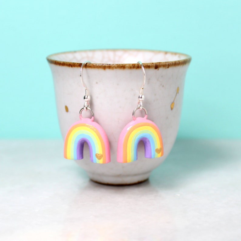 A pair of pastel rainbow arch earrings with tiny golden hearts. The earrings are hanging from a small cup in front of a bright blue background.