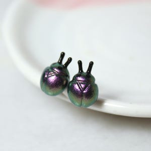 A pair of purple and silver beetle stud earrings rest against a white jewelry dish