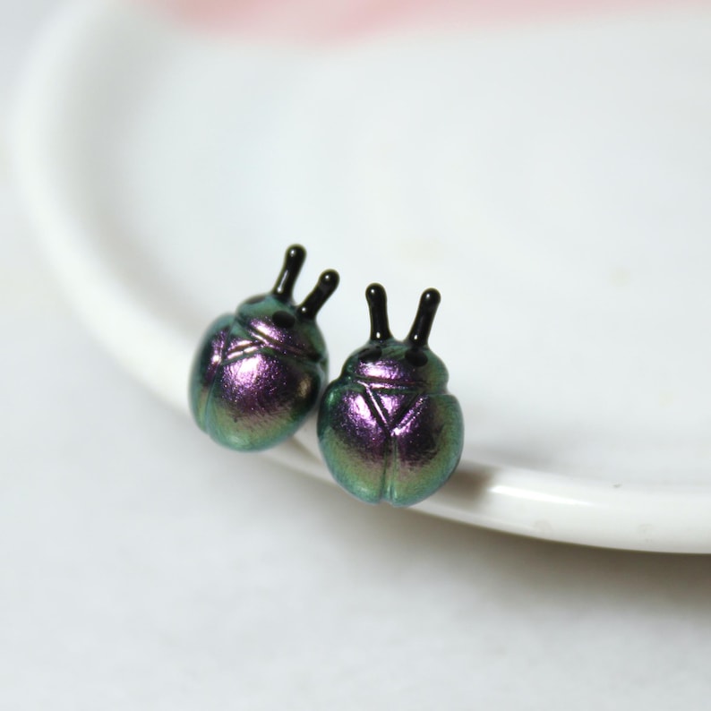 A pair of purple and silver beetle stud earrings rest against a white jewelry dish