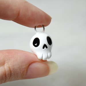 May include: A small, white skull pendant with black eye sockets and a nose. The pendant has a silver-colored loop at the top for attaching to a necklace or keychain. The skull is held between two fingers.