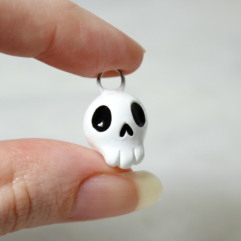 May include: A small, white skull pendant with black eye sockets and a nose. The pendant has a silver-colored loop at the top for attaching to a necklace or keychain. The skull is held between two fingers.