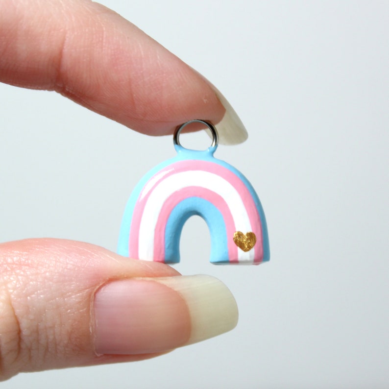 A hand holding a trans pride rainbow charm. The charm is about 3/4 of an inch tall.