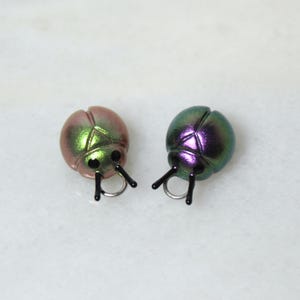 May include: Two iridescent ladybug-shaped charms. One is a blend of pink and green, the other is green and purple. Each has black antennae and a silver ring. These charms are suitable for jewelry making or craft projects.