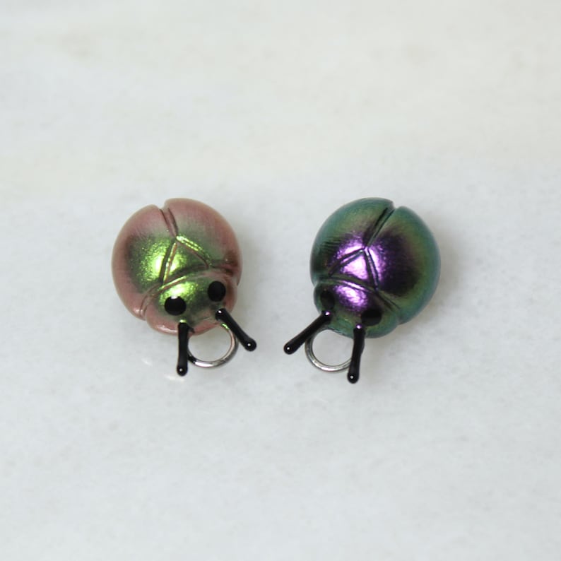 May include: Two iridescent ladybug-shaped charms. One is a blend of pink and green, the other is green and purple. Each has black antennae and a silver ring. These charms are suitable for jewelry making or craft projects.