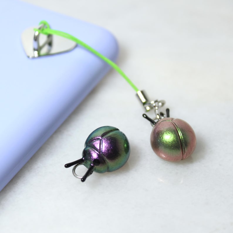 May include: Two iridescent bug-shaped phone charms. One is green and purple, the other is pink and green. They are attached to a green cord and a silver phone grip.