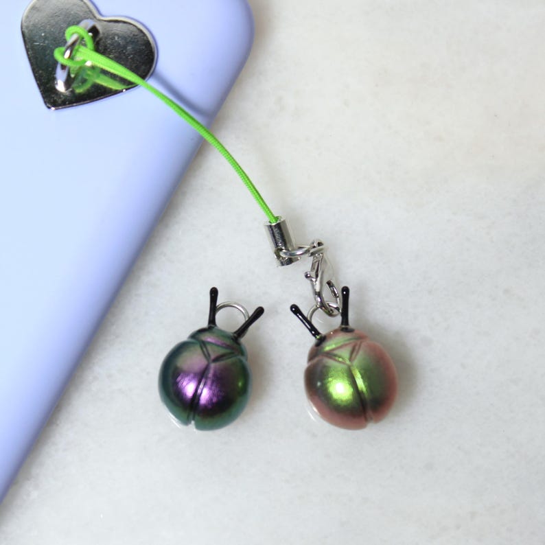 May include: Two iridescent beetle charms with black antennae are attached to a green cord and silver-tone hardware. The charms are a mix of green, pink, and purple. The cord is attached to a light purple phone case with a silver heart-shaped charm.
