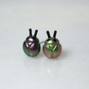 Two different colors of beetle stud earrings sit next to each other: one in purple and silver, and the other in green and gold.