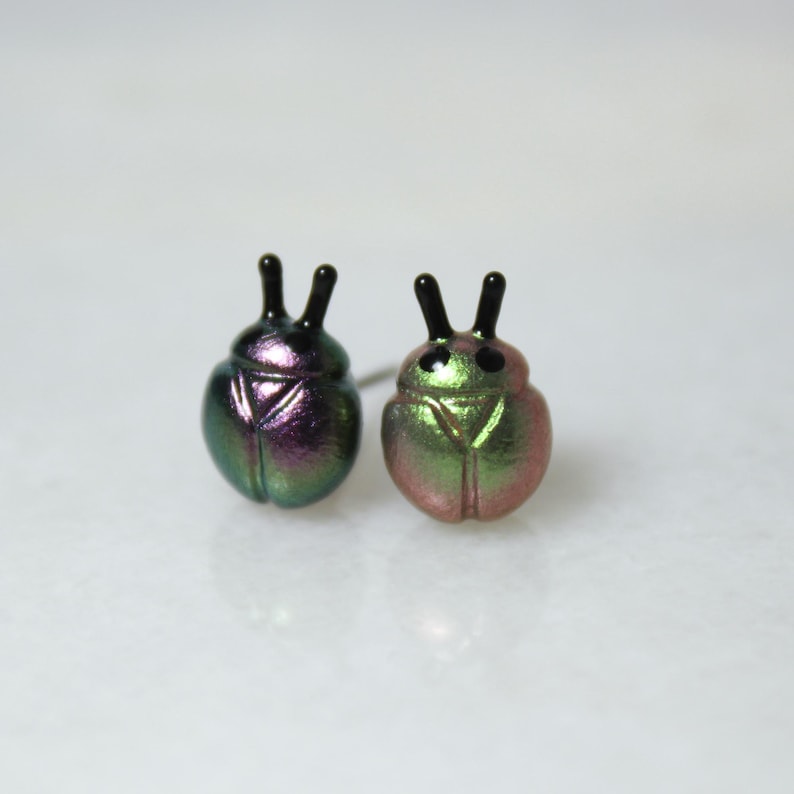 Two different colors of beetle stud earrings sit next to each other: one in purple and silver, and the other in green and gold.