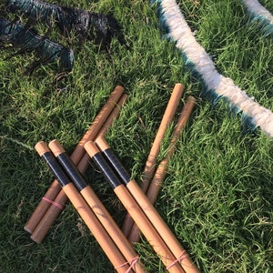 Könnte beinhalten: Mehrere Holzstäbe mit schwarzen Klebebändern sind auf grünem Gras angeordnet. Einige Stäbe sind mit rosa Gummibändern befestigt. Schwarze und weiße Federboas sind ebenfalls zu sehen.