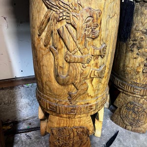 Könnte beinhalten: Eine zylindrische Holztrommel mit detaillierten handgeschnitzten Reliefs einer Katzenfigur und geometrischen Mustern. Die Trommel steht auf Holzbeinen und ist mit einem schwarz-weißen Webstoffband mit Quasten verziert.