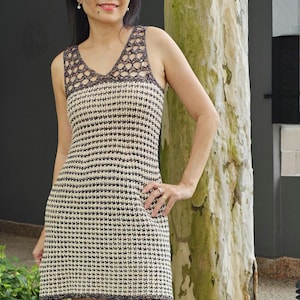 May include: A woman wearing a white and brown crochet dress with a crocheted lace neckline. She is standing in front of a tree trunk.