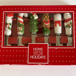 May include: A set of six Christmas-themed cheese knives in a red box with white polka dots. Each knife features a different festive handle design, including a candy cane, Christmas tree, and Santa. The box reads "HOME FOR THE HOLIDAYS".
