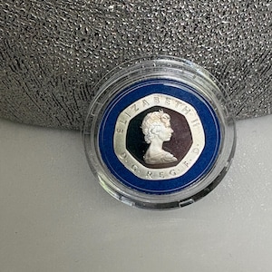 May include: A silver coin encased in a clear plastic holder. The coin features a profile of a person and the text "ELIZABETH II D.G. REG. F.D." around the edge. The coin is set against a blue background.