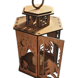 May include: A wooden lantern with a Bigfoot theme. The lantern is made of light brown wood and features cut-out designs of Bigfoot, trees, a moon, and stars. It has a handle on top and a door that opens to reveal a candle.