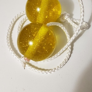 May include: Two translucent yellow spherical balls with embedded gold glitter, connected by a white braided nylon cord. The spheres are smooth and glossy, featuring a central internal channel through which the white rope is threaded and knotted.