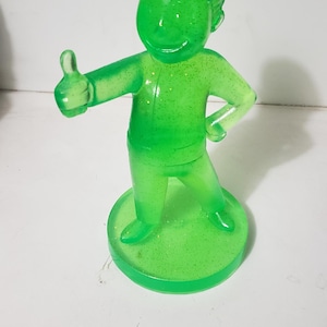 May include: A translucent neon green figurine of a character with a stylized hairstyle, wearing a collared shirt and pants, standing on a circular base. The figure is posed with one arm extended, giving a thumbs-up gesture, and the other hand on a hip.