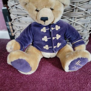 Puede incluir: Un oso de peluche beige con una chaqueta de terciopelo morado con detalles dorados. El oso tiene almohadillas moradas y la palabra "Harrods" y el año "2004" bordados en una pata. El oso está sentado sobre una superficie burdeos.