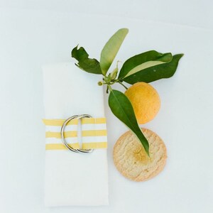 May include: A white envelope tied with a yellow and white striped ribbon and a silver buckle. A lemon and a lemon cookie are on the white surface.
