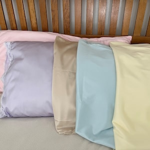 May include: A collection of satin pillowcases in various pastel colors, including pink, lavender, beige, light blue, and yellow. The pink pillowcase has a lace trim. The pillowcases are arranged on a bed.