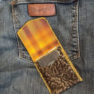 Peut inclure: Un portefeuille en cuir marron avec un rabat en tissu écossais jaune et une poche en cuir marron à motif floral. Le portefeuille est dans la poche arrière d'un jean bleu avec une étiquette Wrangler en cuir marron.