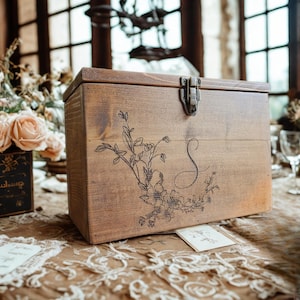 May include: A light brown wooden box with a hinged lid and metal clasp. The box is decorated with floral and vine etchings and a cursive letter "S". The box sits on a lace-covered table, with flowers in the background.