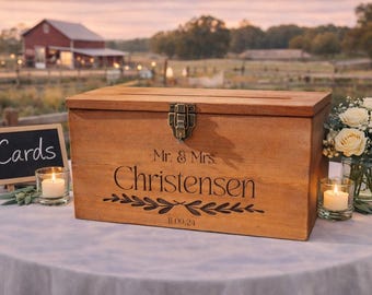 Peut inclure: Une boîte rectangulaire en bois avec un couvercle à charnière et un loquet en métal. L'avant est gravé avec le texte Mr. & Mrs. Christensen, un motif de feuilles décoratif et la date 11.09.24. Une fente est découpée sur le couvercle.