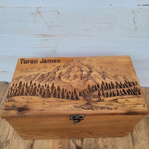 May include: A rectangular wooden box with a hinged lid featuring a laser-engraved mountain landscape, a forest of pine trees, and the name Toran James. The box has a natural wood grain finish and a small, dark metal latch on the front center.
