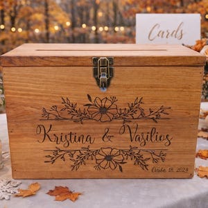 May include: A wooden card box with a hinged lid and metal clasp. The box is engraved with floral designs and the names "Kristina & Vasilios" along with the date "October 18, 2024".