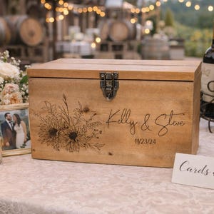 May include: A wooden card and gift box with a floral design and the names "Kelly & Steve" and the date "11/23/24" engraved on the front. A metal clasp secures the lid. A sign reads "Cards & Gifts".