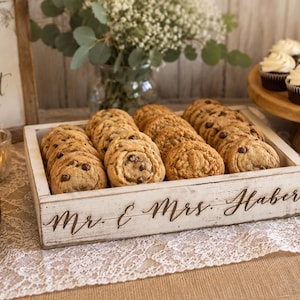 May include: A rectangular, distressed white wooden tray filled with chocolate chip cookies. The front side of the tray features the engraved text "Mr. & Mrs. Haber" in a cursive script font. The tray has a rustic finish with visible wood grain.