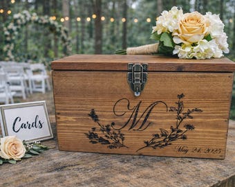 Peut inclure: Une boîte rectangulaire en bois avec un loquet en métal, ornée d'une lettre M gravée en noir entourée d'illustrations florales et du texte May 24, 2025. Un bouquet de roses pêche et blanches est posé dessus, avec un panneau Cards.