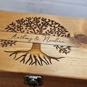 May include: A rectangular wooden box with a hinged lid featuring a laser-engraved tree of life design. The center of the tree includes a horizontal banner with the text Anthony & Nicolina. A small metal latch is visible on the front edge of the box.