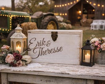 Peut inclure: Une boîte rectangulaire en bois blanc avec un loquet en métal, portant l'inscription en lettres cursives marron foncé Erik & Therese. Elle est présentée avec une lanterne blanche, une noire, des fleurs et des bougies sur une table.