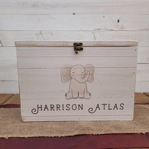May include: A rectangular white wooden storage box with a hinged lid and a bronze-coloured metal latch. The front features a printed illustration of a light brown elephant and the black text "HARRISON ATLAS" in a decorative serif font.